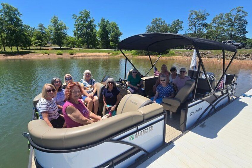 A group of people on a pontoon boat with a canopy, docked at a lake with trees in the background. 5
