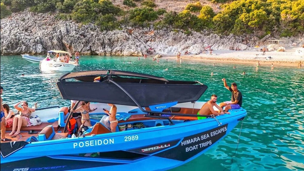 Ein blaues Boot namens Poseidon 2989 auf ruhigem Wasser mit einer felsigen Küste und Menschen, die im Hintergrund schwimmen. 5