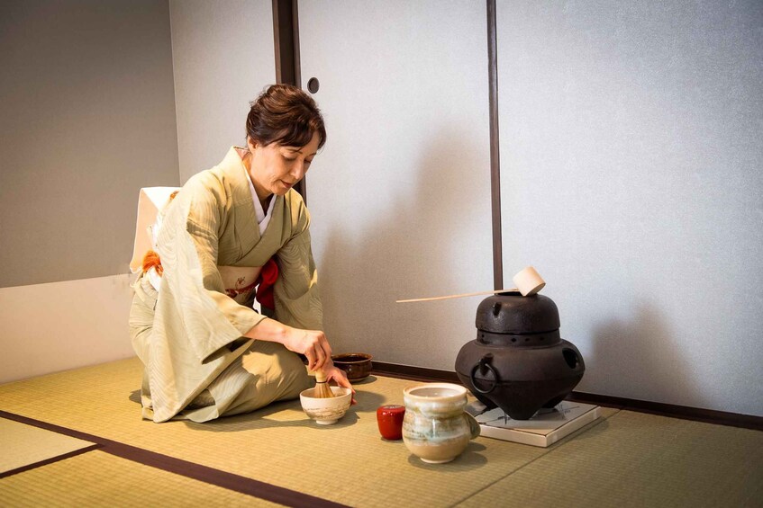 A person in traditional attire performing a tea ceremony with a tea pot and bowls on tatami mat flooring. 1