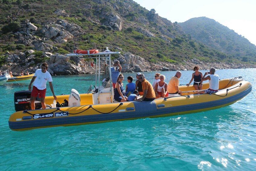 A group of people on a yellow inflatable boat with a Suzuki outboard motor, surrounded by calm blue water and rocky hills in the background. 5