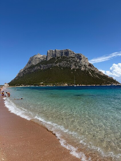 A beach with clear turquoise water, a rocky mountain in the background, and a few people in the water. 3