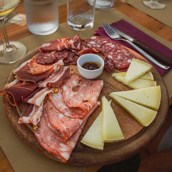 A wooden board with various cured meats and cheese slices, accompanied by a small bowl of sauce and a glass of water. 2