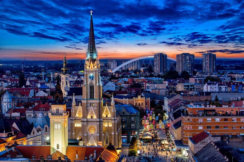 A cityscape at dusk with a prominent church featuring a tall spire and clock tower, surrounded by residential buildings and a modern bridge. 5