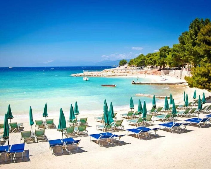 A beach with rows of sun loungers and umbrellas, clear turquoise water, and a rocky coastline with trees. 1