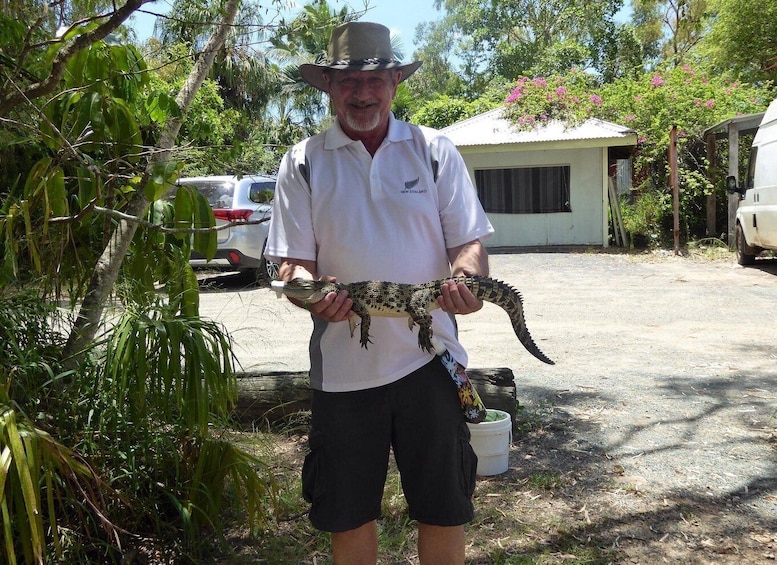 A person holding a large reptile, possibly a crocodile, outdoors with trees and a building in the background. 3