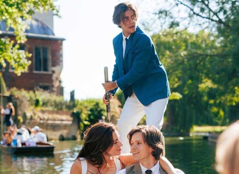 A person in a blue shirt and white pants standing on a boat, holding a pole, with a river and buildings in the background. 1