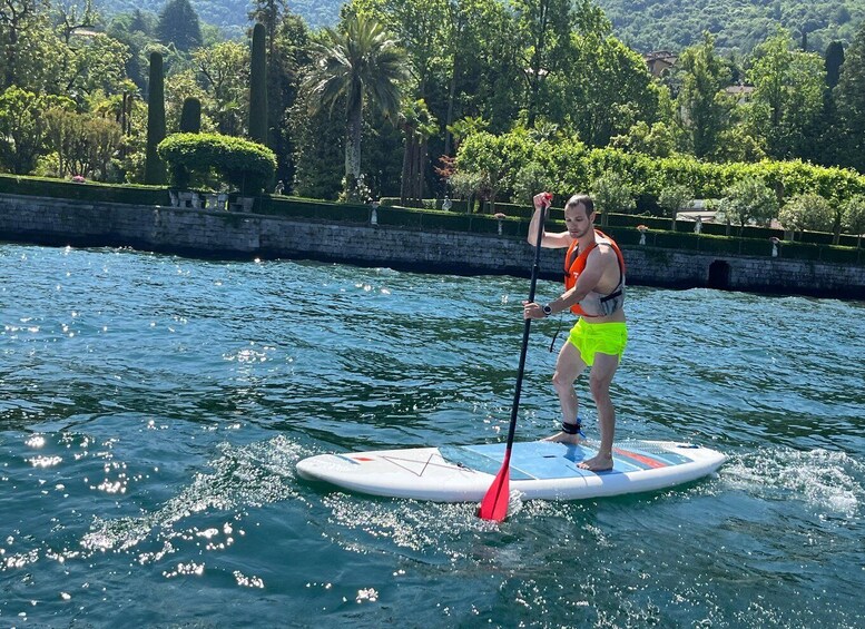 A person paddleboarding on a calm body of water with greenery and buildings in the background. 1