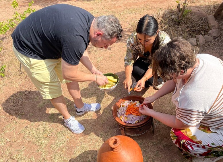 Three people preparing food outdoors with vegetables and a clay pot. 3