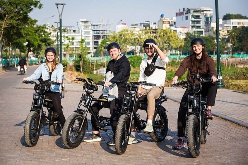 Four people riding electric bicycles on a paved path with buildings and trees in the background. 1