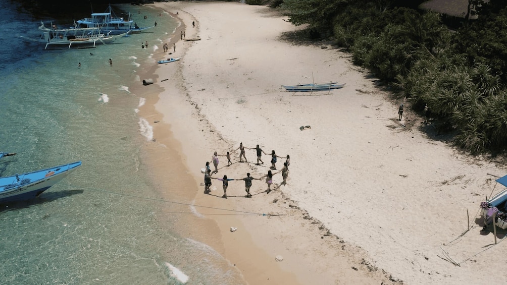 A sandy beach with people holding hands in a circle, boats in the water, and dense vegetation on the right. 2