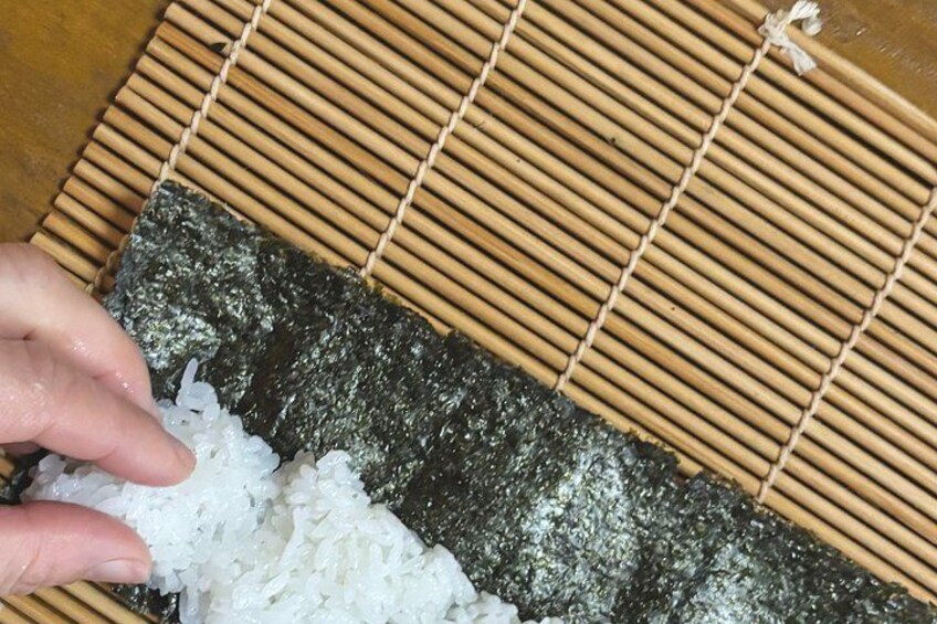 A hand placing white rice on a sheet of nori (seaweed) on a bamboo mat. 4