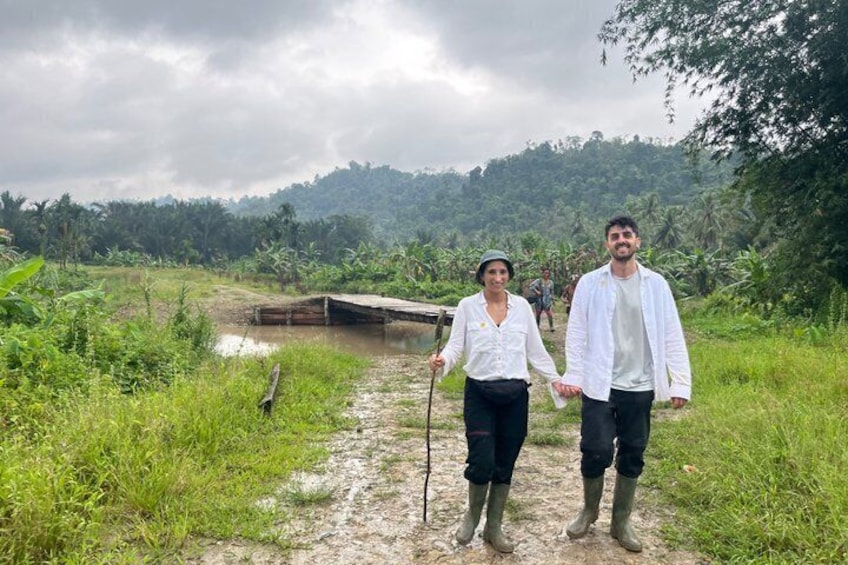 Two people walking on a path with a wooden bridge over a water body, surrounded by greenery and hills. 3