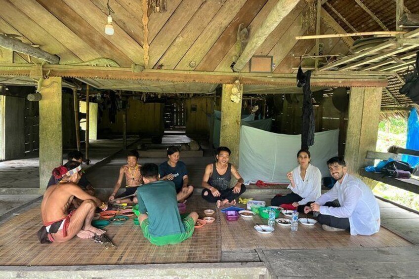 A group of people sitting on the floor in a wooden structure with a thatched roof. 5