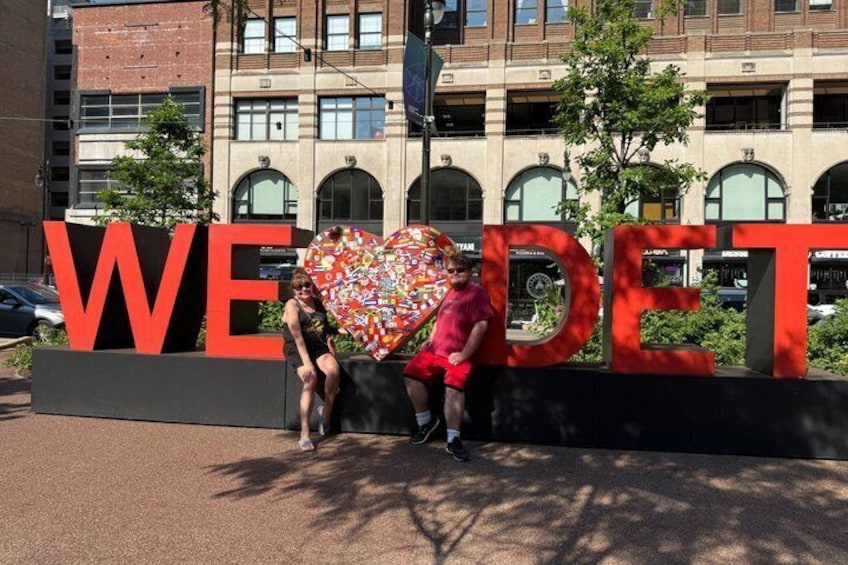 A couple sitting in front of a large sign with the word 'WEDELT' and a heart-shaped decoration. 5