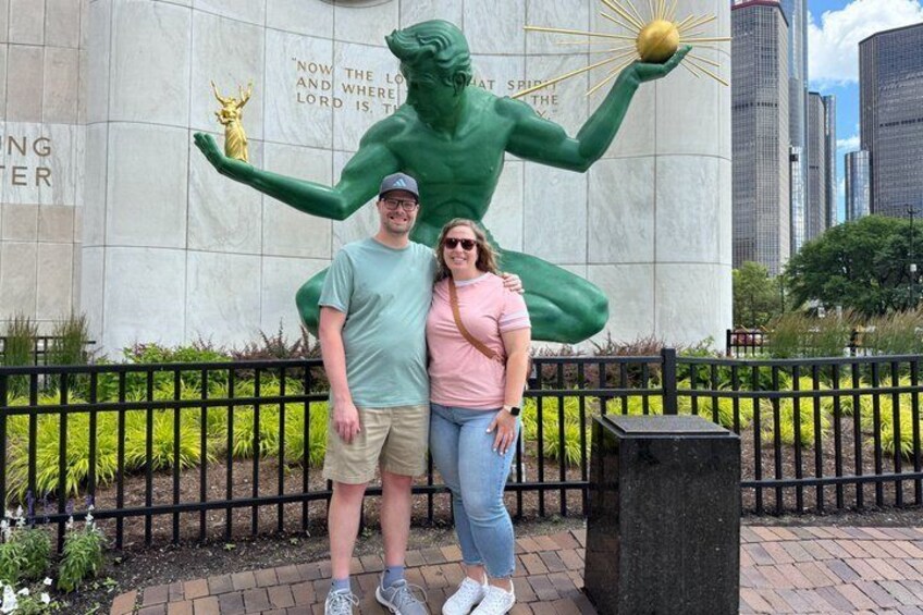 A couple standing in front of a large green statue with a golden sphere, with a cityscape in the background. 1