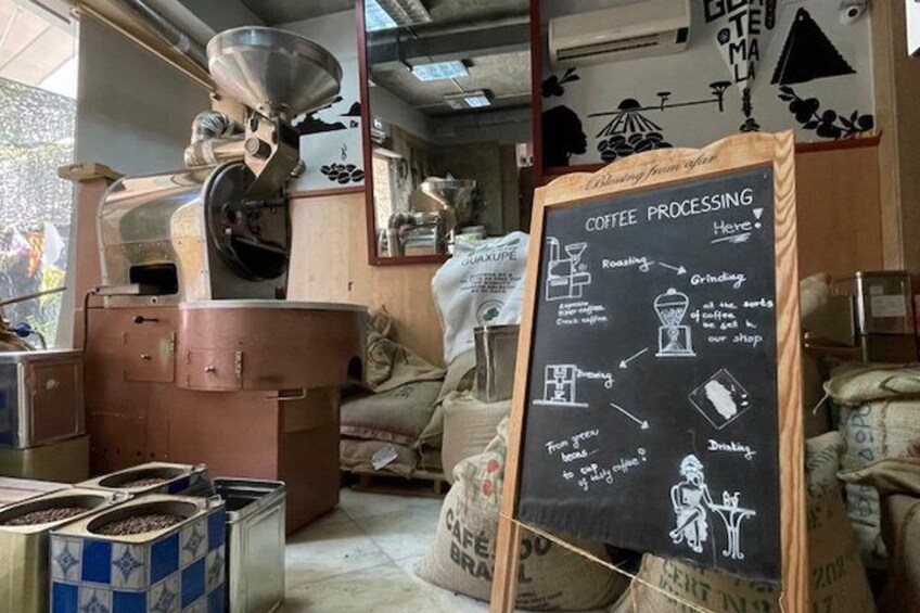 A coffee processing area with a chalkboard explaining the steps, coffee beans in bins, and a large coffee roaster. 1