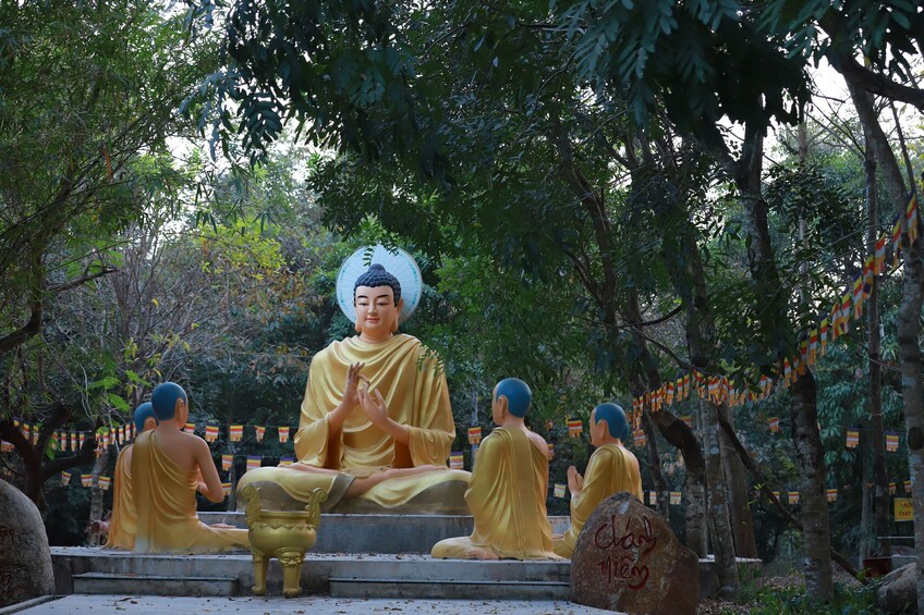 Eine Statue einer sitzenden Figur in Meditationshaltung, umgeben von Bäumen und einem Stein mit Inschriften. 3