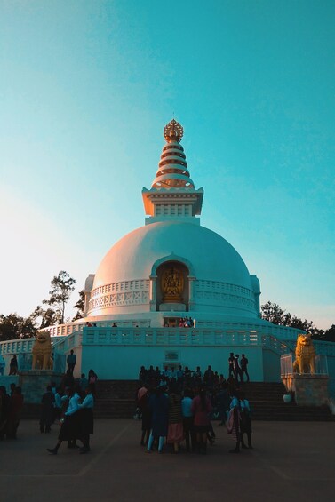 Eine große weiße Stupa mit einer goldenen Statue am Fuß, umgeben von einer Menschenmenge. 4