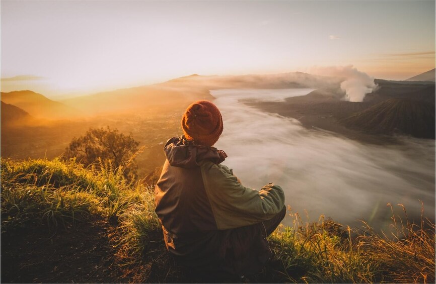 A person sitting on a grassy hill overlooking a valley with a mountain in the distance. 1