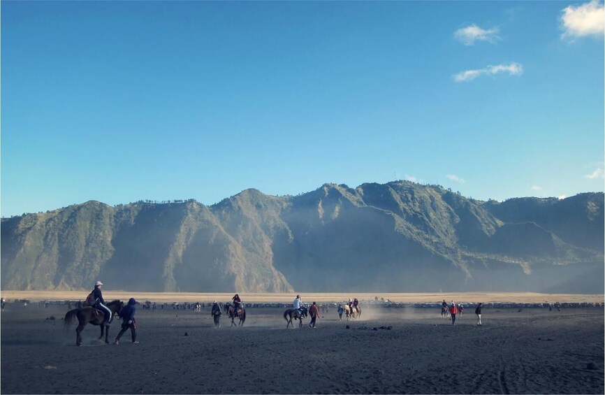 A group of people riding horses on a sandy beach with mountains in the background. 3