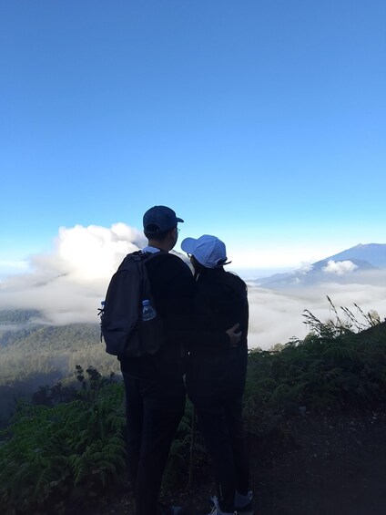 Two people standing on a mountain trail, overlooking a valley with clouds below and a mountain in the distance. 2