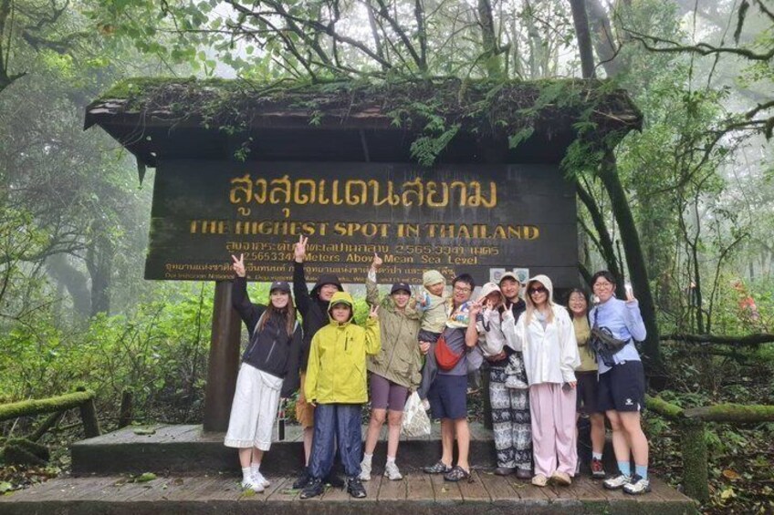 A group of people standing in front of a sign that reads 'THE HIGHEST SPOT IN THAILAND' with a forested background. 4