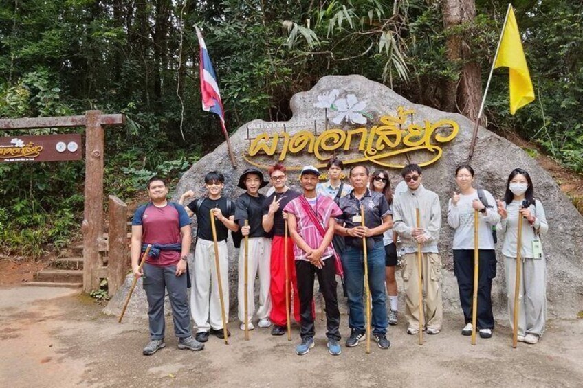 A group of people standing in front of a large rock with inscriptions, holding walking sticks, and a signboard with flags. 5