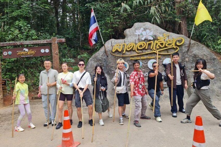 A group of people posing in front of a sign with Thai script, surrounded by trees and a flag. 3