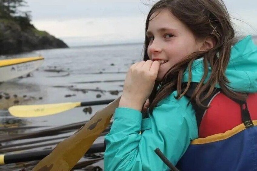 A person in a teal jacket holding a red backpack, with a row of yellow paddles and a boat in the background. 5