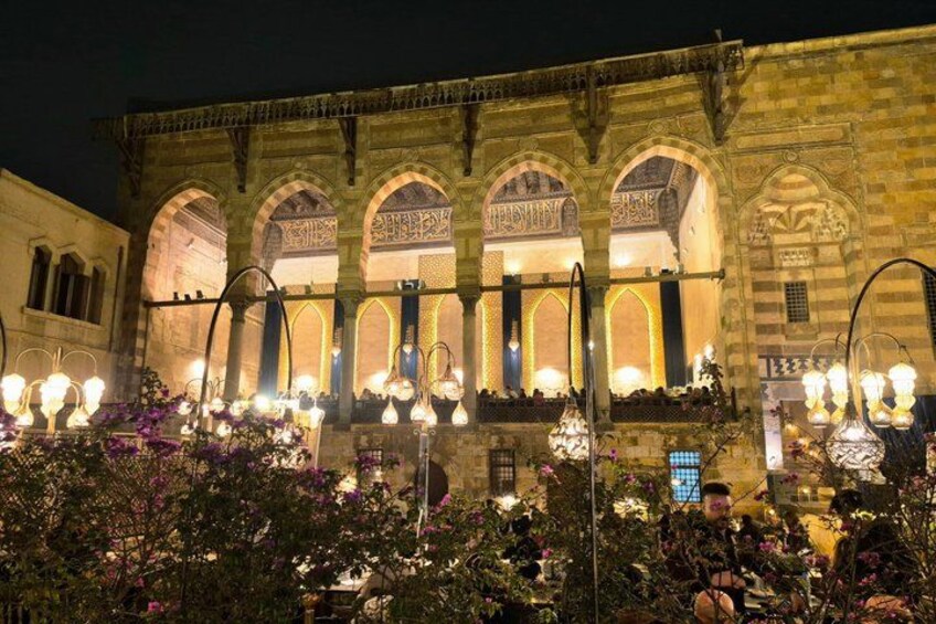 A historic building with arched windows and intricate carvings, illuminated by street lamps and surrounded by flowering plants. 2