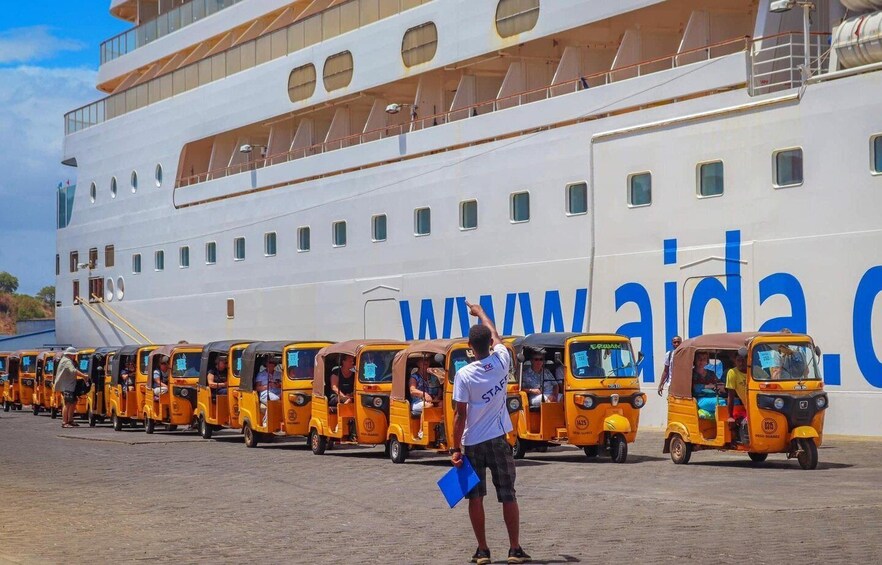A large cruise ship with multiple yellow auto rickshaws lined up in front. 1