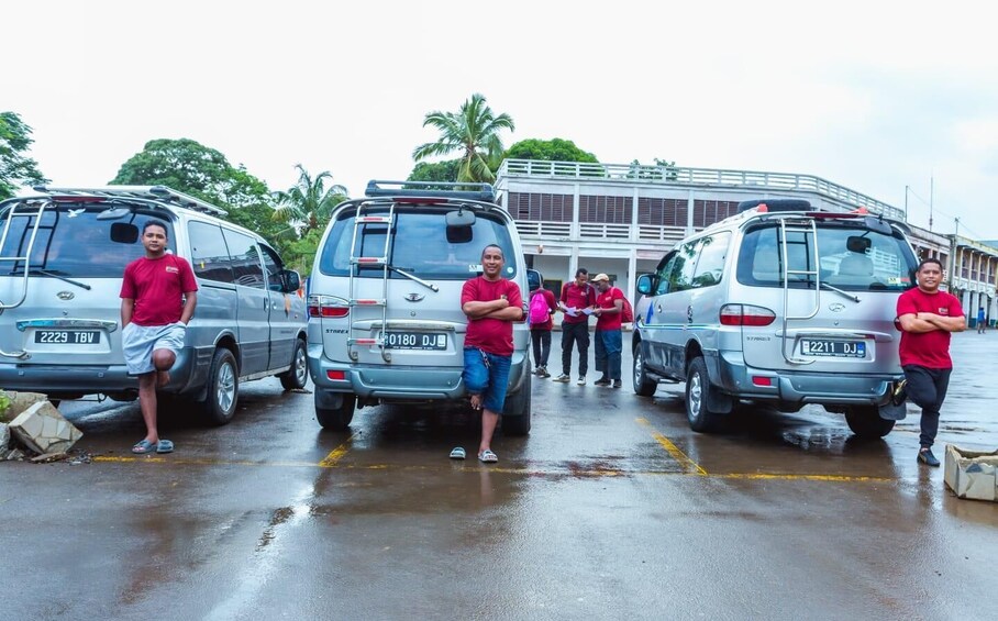 Three people standing in front of three silver vans parked on a wet road with palm trees in the background. 4