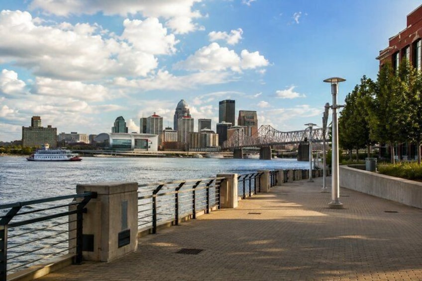 A riverside walkway with a city skyline, a bridge, and a boat on the water. 5