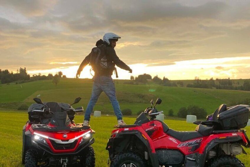 A person standing between two red quad bikes in a grassy field with a sunset in the background. 3