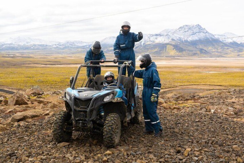 Three people in blue protective suits operating an ATV on a rocky terrain with a mountainous backdrop. 2
