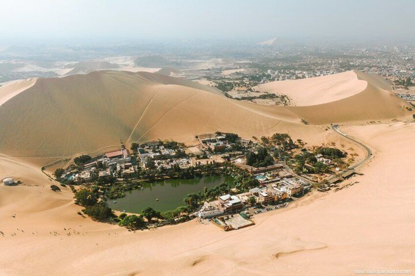 A desert landscape with sand dunes and a small settlement with buildings and a pond. 4