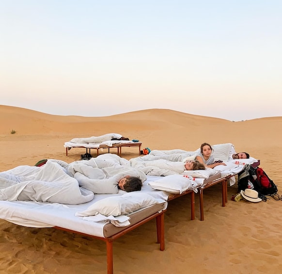 Four people lying on beds in a desert with sand dunes in the background. 2