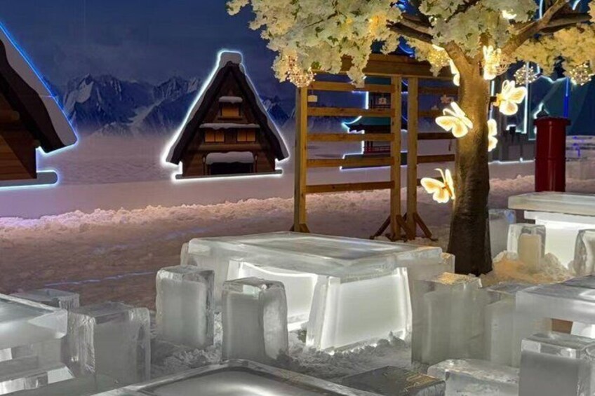 An outdoor dining area with ice tables and chairs, a wooden structure resembling a house, and a tree with illuminated flowers. 2