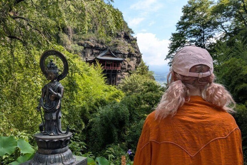 A person in an orange garment and pink cap, a statue, and a traditional building in a lush green setting. 1