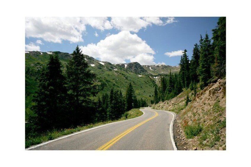 A winding mountain road with a double yellow line, surrounded by dense green forests and snow-capped peaks under a partly cloudy sky. 3
