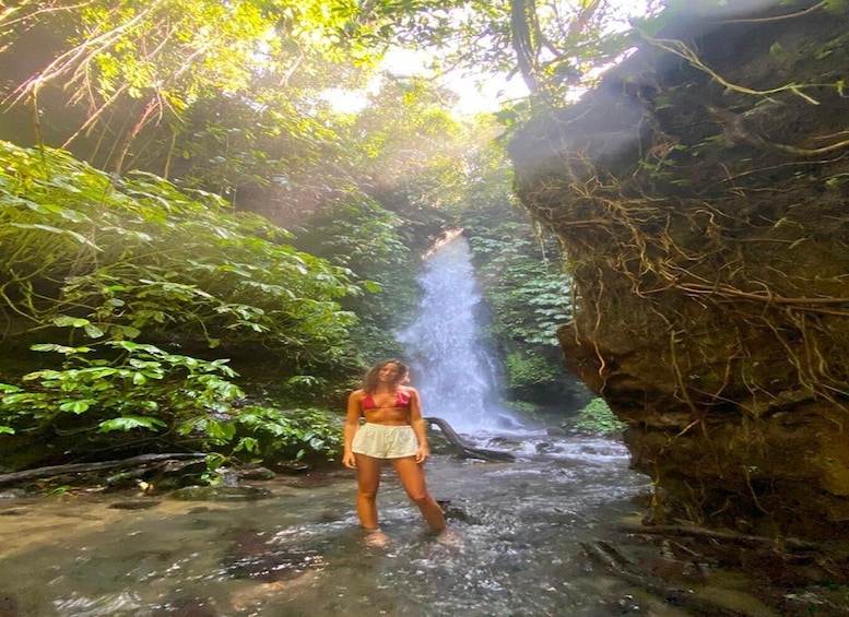 A person standing in a shallow stream surrounded by lush greenery and a waterfall in the background. 3