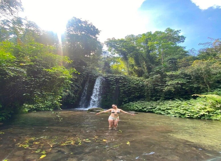 A person standing in a shallow stream with a waterfall in the background surrounded by dense greenery. 5