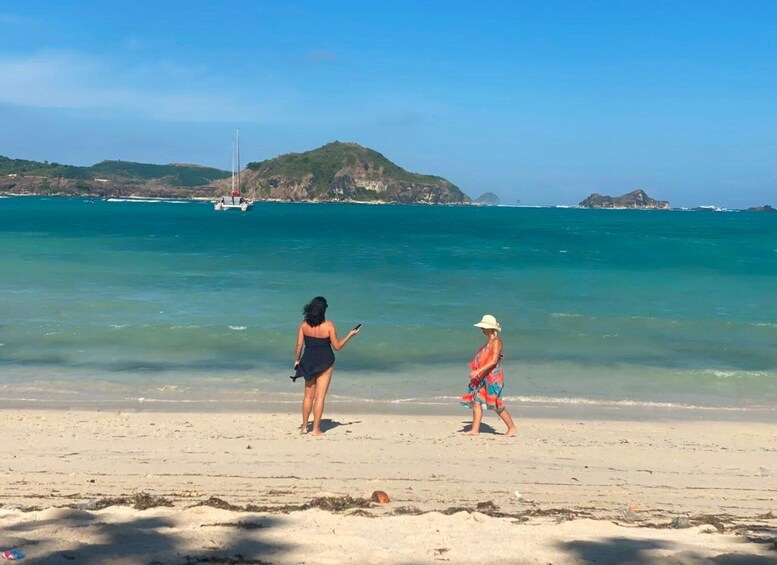 A beach with two people walking, a boat in the water, and a hilly landscape in the background. 4