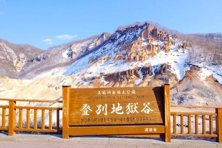 A wooden sign with text in a foreign language, a mountainous landscape with snow, and a wooden fence. 1