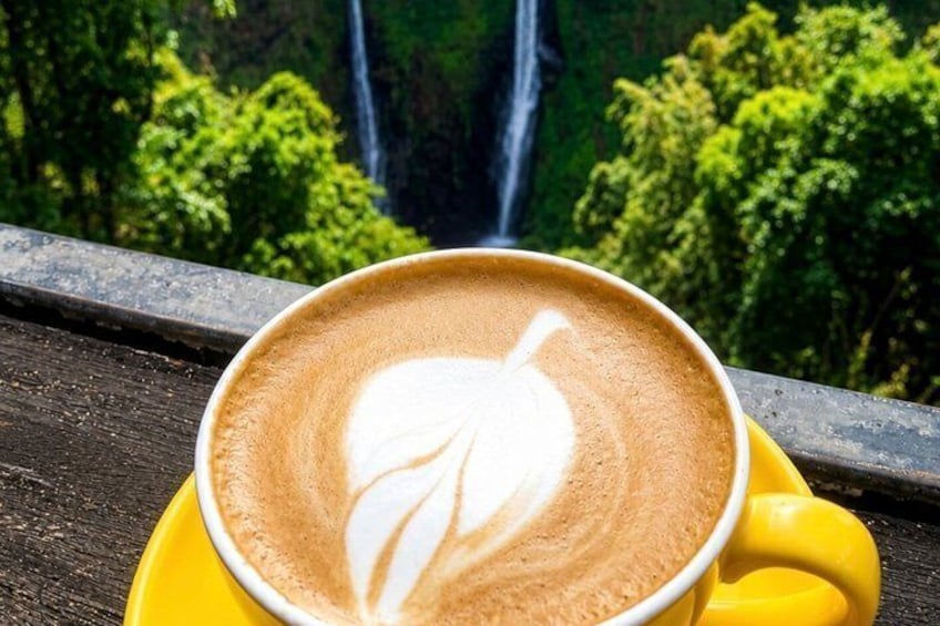 A cup of coffee with latte art on a yellow saucer, set against a backdrop of a waterfall and lush greenery. 1