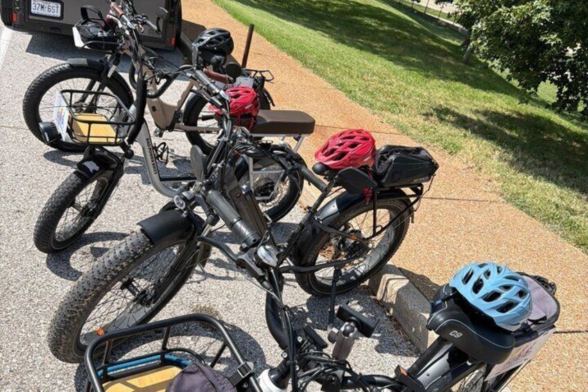 A row of bicycles parked on a sidewalk with helmets and bags attached. 2
