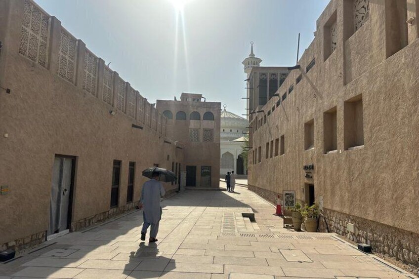 A sunlit courtyard with a person walking, beige buildings with arched windows, and a distant minaret. 4