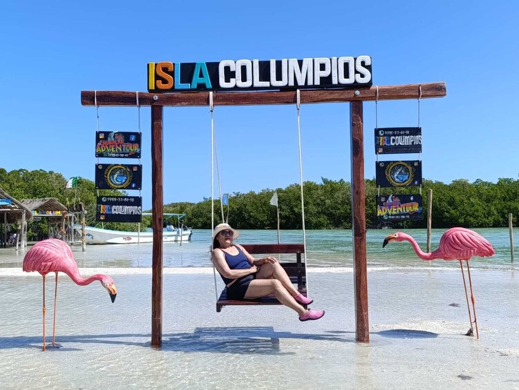 A woman sitting on a swing at Isla Columpios, with flamingos and a boat in the background. 2