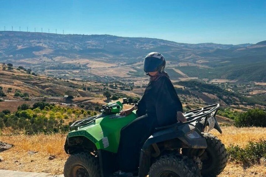 A person riding a green ATV on a hillside with a view of rolling hills and wind turbines in the distance. 3