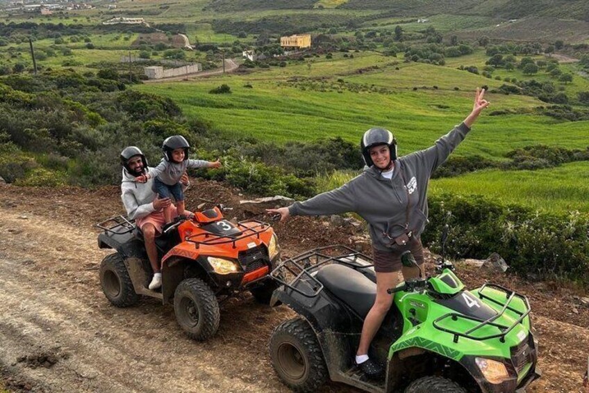 Three people on ATVs on a dirt path with green fields and a building in the background. 1